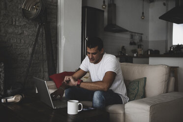 Young man sitting on sofa at home using laptop and checking the time - ERRF00357