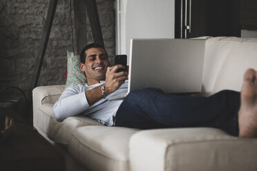 Happy young man lying on sofa at home using cell phone and laptop - ERRF00359