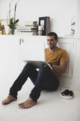 Lächelnder junger Mann, der auf dem Boden sitzt und einen Laptop benutzt, lizenzfreies Stockfoto