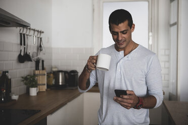 Lächelnder junger Mann mit einer Tasse Kaffee und einem Mobiltelefon in der Küche zu Hause - ERRF00369