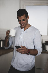Lächelnder junger Mann mit einer Tasse Kaffee und einem Mobiltelefon in der Küche zu Hause - ERRF00370