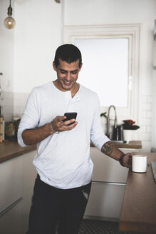 Lächelnder junger Mann mit einer Tasse Kaffee und einem Mobiltelefon in der Küche zu Hause - ERRF00371