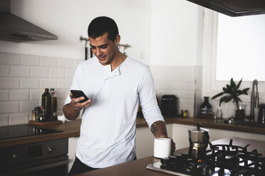 Lächelnder junger Mann mit einer Tasse Kaffee und einem Mobiltelefon in der Küche zu Hause - ERRF00372