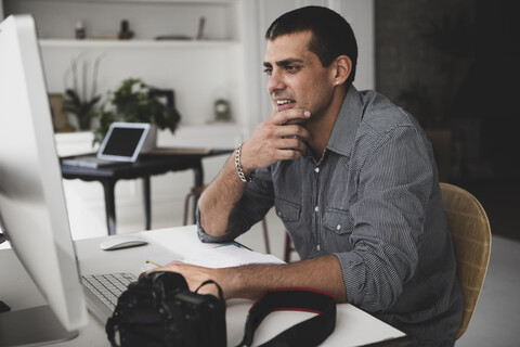 Junger Mann mit Kamera, der am Schreibtisch sitzt und einen Computer benutzt, lizenzfreies Stockfoto