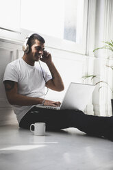Lächelnder junger Mann mit Kopfhörern, der auf dem Boden sitzt und einen Laptop benutzt - ERRF00385