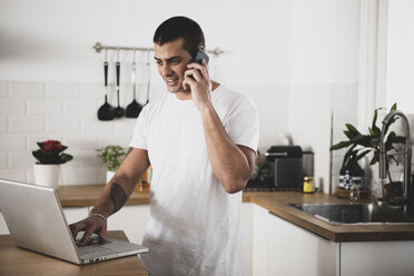 Lächelnder junger Mann mit Laptop und Handy in der Küche zu Hause - ERRF00390