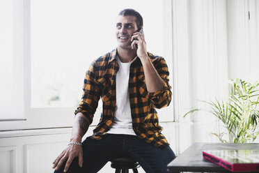 Lächelnder junger Mann, der am Fenster mit seinem Handy telefoniert - ERRF00393