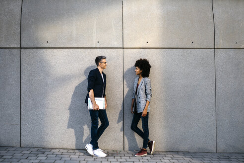 Two colleagues talking outdoors leaning against a wall - JRFF02192