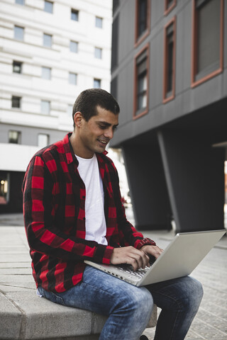 Junger Mann sitzt im Freien in der Stadt und benutzt einen Laptop, lizenzfreies Stockfoto