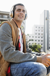 Lächelnder junger Mann, der in der Stadt im Freien sitzt und Kopfhörer trägt - ERRF00404