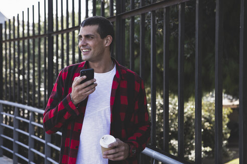 Lächelnder junger Mann mit Kaffee zum Mitnehmen und Mobiltelefon unterwegs - ERRF00408