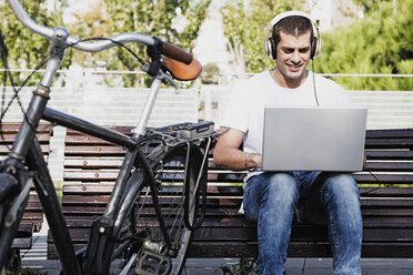 Young man sitting on a bench wearing headphones and using laptop next to bicycle - ERRF00411