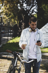 Young man with bicycle and takeaway coffee checking cell phone - ERRF00415