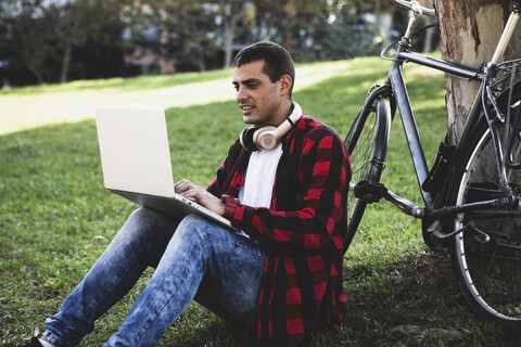 Junger Mann sitzt in einem Park und benutzt einen Laptop neben einem Fahrrad, lizenzfreies Stockfoto