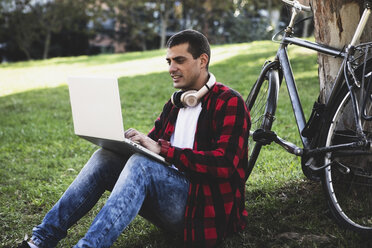 Junger Mann sitzt in einem Park und benutzt einen Laptop neben einem Fahrrad - ERRF00416