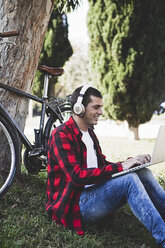 Junger Mann sitzt in einem Park, trägt Kopfhörer und benutzt einen Laptop neben einem Fahrrad - ERRF00417