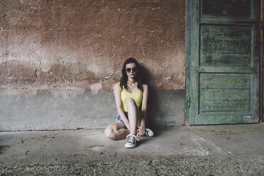 Porträt einer Frau mit Sonnenbrille, die an einer alten Mauer sitzt - CAVF60481