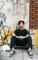 Porträt eines ernsten Mannes, der auf einem Skateboard an einer Wand in der Stadt sitzt - CAVF60485