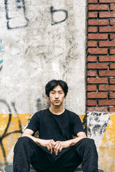 Portrait of man sitting on skateboard against wall in city - CAVF60486