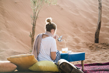 Rückansicht einer Frau, die in der Wüste auf einer Decke sitzend etwas trinkt - CAVF60501