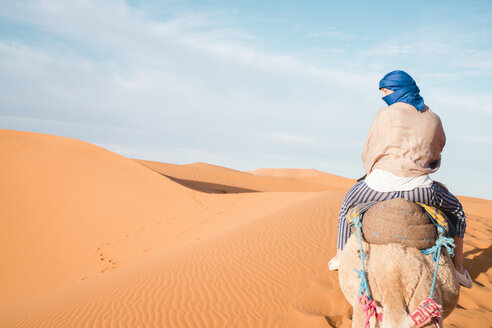 Rückansicht einer Frau mit Kopftuch auf einem Kamel in der Wüste von Merzouga gegen den Himmel - CAVF60504