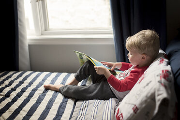 Hochformatiger Blick auf einen Jungen, der ein Bilderbuch hält, während er zu Hause auf dem Bett sitzt - CAVF60512