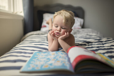 Junge mit Händen am Kinn liest Bilderbuch, während er zu Hause auf dem Bett liegt - CAVF60514