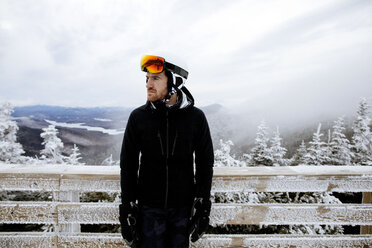 Thoughtful man wearing ski-wear while standing against frozen wooden fence during foggy weather - CAVF60521