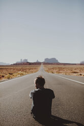 Rear view of hiker sitting on country road amidst desert against clear sky during sunny day - CAVF60539