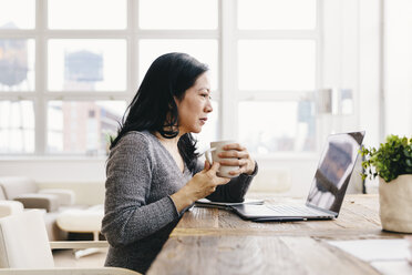 Geschäftsfrau hält eine Kaffeetasse, während sie auf einen Laptop-Computer auf einem Schreibtisch im Büro schaut - CAVF60571