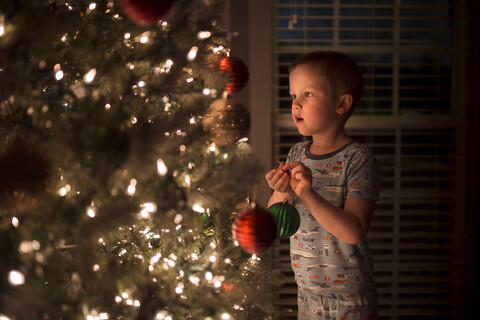 Junge hält eine Weihnachtskugel und betrachtet einen beleuchteten Weihnachtsbaum, lizenzfreies Stockfoto
