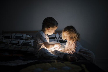 Siblings with illuminated string lights sitting on bed at home - CAVF60605