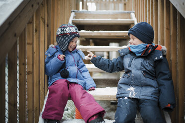 Glückliche Geschwister, die im Winter auf einer Treppe sitzend mit Schnee spielen - CAVF60608