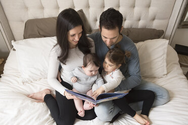 Hochformatige Ansicht von Eltern mit Kindern, die auf dem Bett sitzend ein Bilderbuch betrachten - CAVF60618
