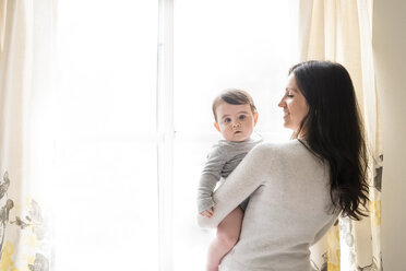 Porträt eines niedlichen Sohnes, der von seiner Mutter gegen ein Fenster getragen wird - CAVF60619