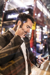 UK, London, businessman with cell phone and earbuds at the bus station by night - WPEF01193