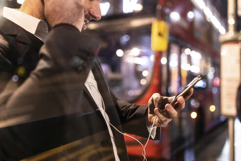 UK, London, Geschäftsmann mit Mobiltelefon und Ohrstöpseln am nächtlichen Busbahnhof - WPEF01194