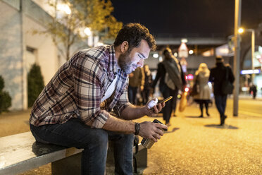 UK, London, lächelnder Mann, der auf einer Bank sitzt und auf sein Handy schaut, bei Nacht - WPEF01204