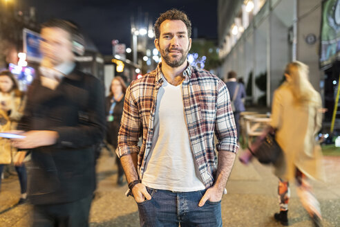 UK, London, portrait of a smiling commuter by night with blurred people passing nearby - WPEF01206