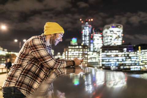 UK, London, smiling man leaning on a railing and looking at his phone with city lights in background stock photo