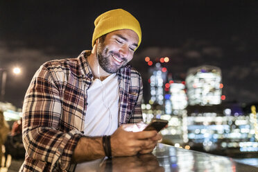 UK, London, lächelnder Mann, der sich auf ein Geländer stützt und auf sein Telefon schaut, mit Stadtlichtern im Hintergrund - WPEF01217
