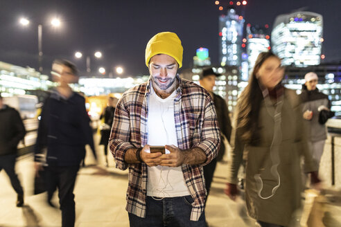 UK, London, smiling man looking at his phone by night with blurred people passing nearby - WPEF01218