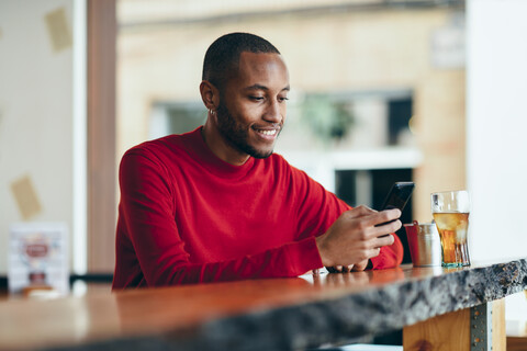Smiling young man wearing red pullover at counter of a bar with soft drink and mobile phone stock photo
