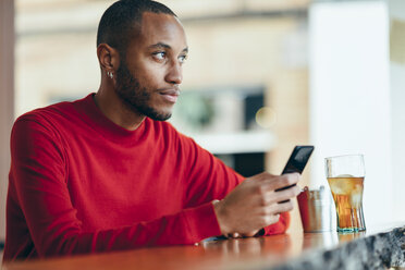 Pensive young man wearing red pullover at counter of a bar with soft drink and mobile phone - JSMF00698