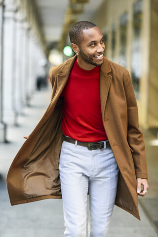 Fashionable young man wearing red pullover and brown coat walking along arcade stock photo