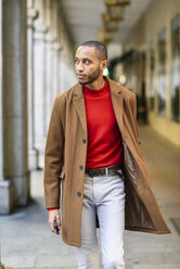 Modischer junger Mann mit rotem Pullover und braunem Mantel, der durch eine Passage geht - JSMF00706