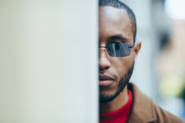 Portrait of bearded young man with nose piercing and sunglasses hiding behind wall - JSMF00707