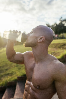 Muskulöser Mann mit nacktem Oberkörper, der eine Pause vom Sport macht und Wasser trinkt - MGOF03870