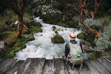 Peru, Huaraz, Man with woolly hat and backpack sitting on wooden bridge at a river - GEMF02694