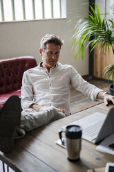 Businessman sitting with feet on desk looking at laptop - GIOF05065